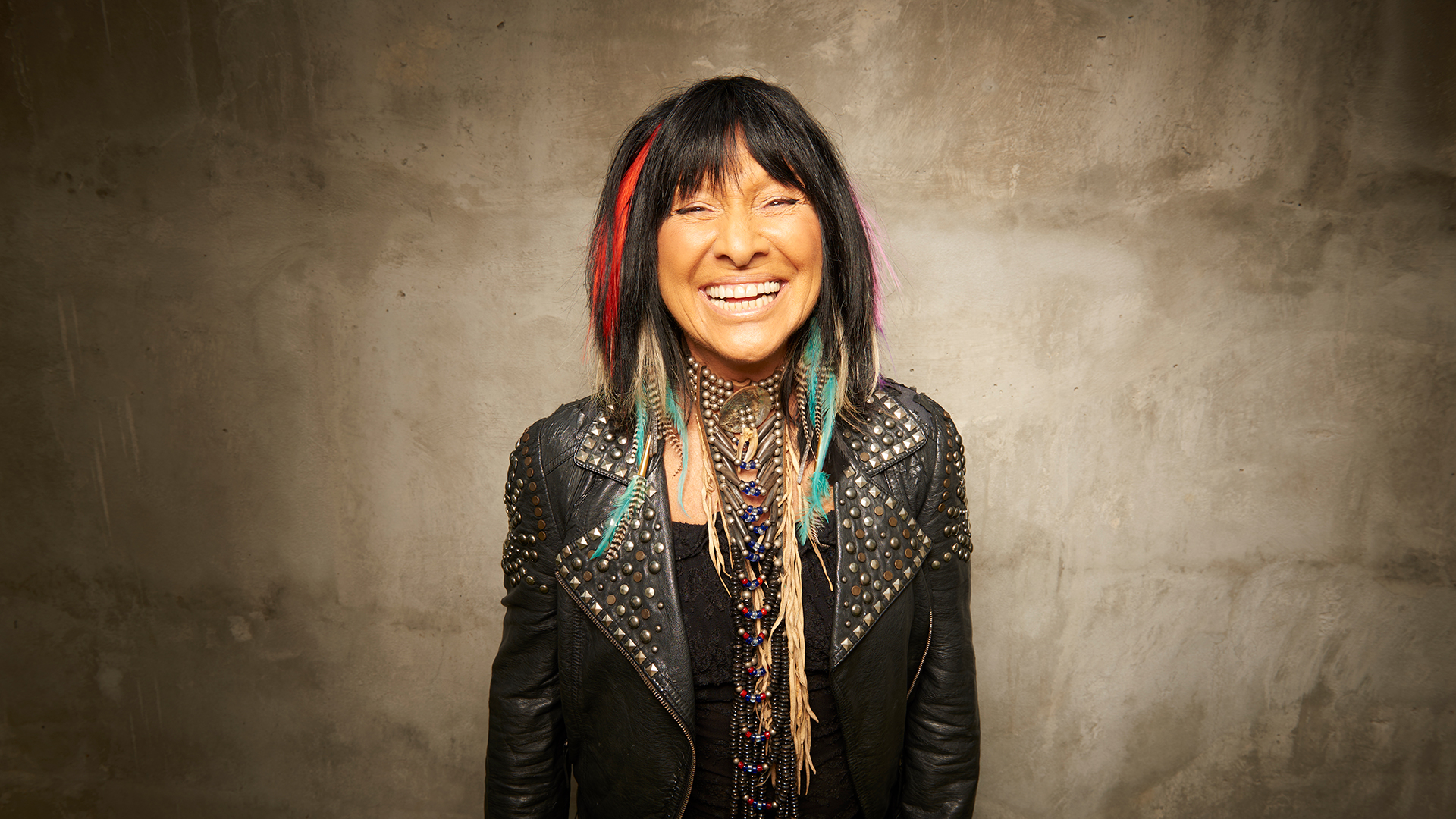 Portrait photograph of Buffy Sainte-Marie wearing a leather jacket, feathers in her hair, and blue jewelry. Brown/dusty backdrop. 