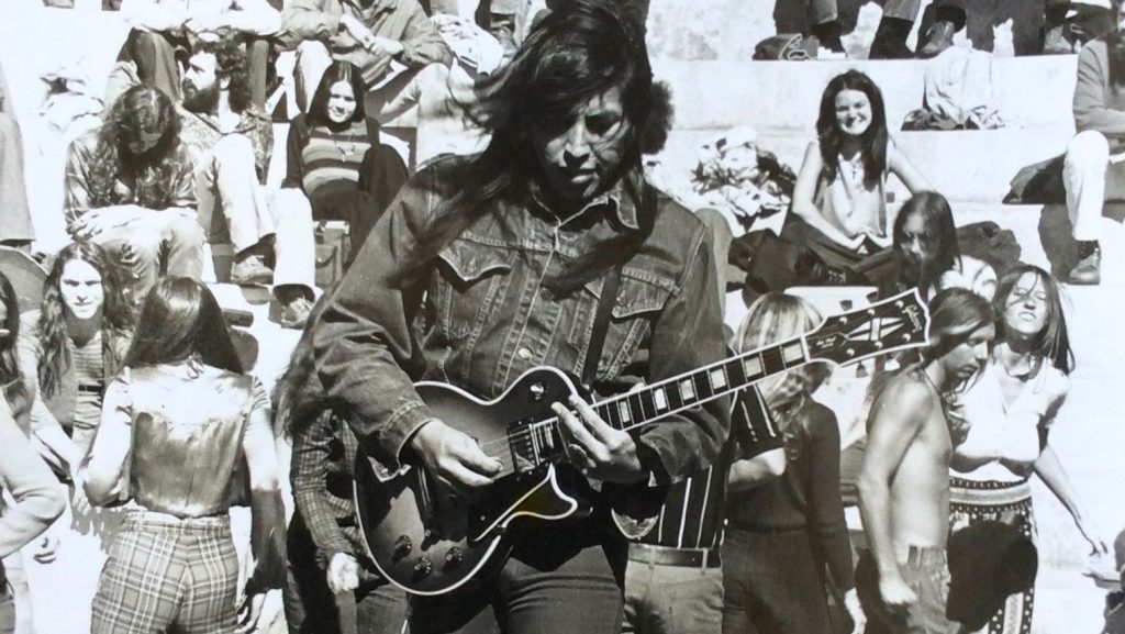 Black and white image of Jesse Ed Davis performing with the view of a crowd behind him with his guitar in hand. 