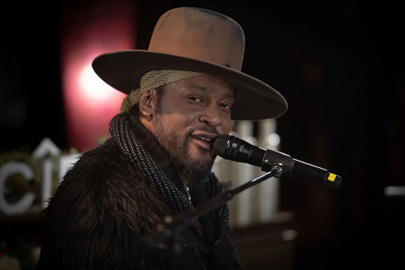 Portrait photograph D'Angelo on stage performing into a microphone. Wearing hat and coat with red background