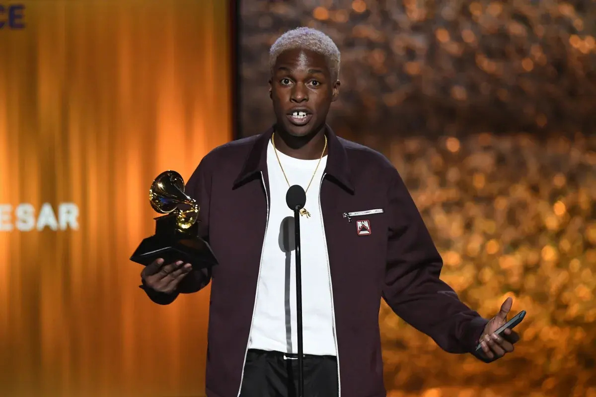 Daniel Caesar on stage expecting an award in a suit, award in hand, and orange background