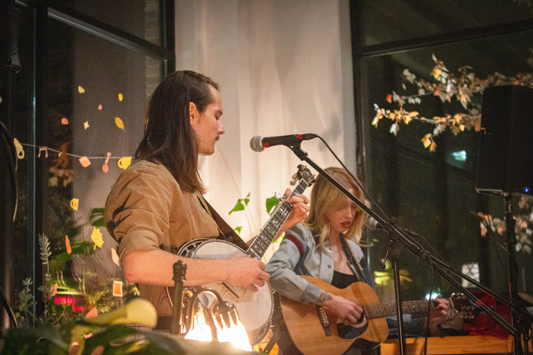 Opener, Babydog, captured in a worms-eye view angle with paper cut leafs in the background. Area lit by lamps. Man closer to camera with tan shirt and guitar, blonde woman with guitar in the back of the shot. 