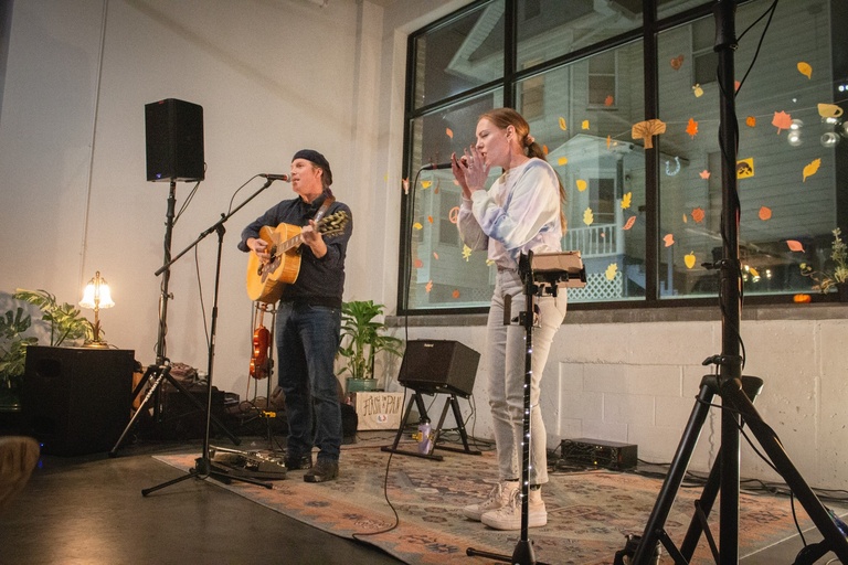 Performers, River Glen, on stage, shot from a worms-eye view. Lady wearing jeans and a blue top holding a microphone. Man wearing jeans and dark long sleeve with a hat holding a guitar and singing. 