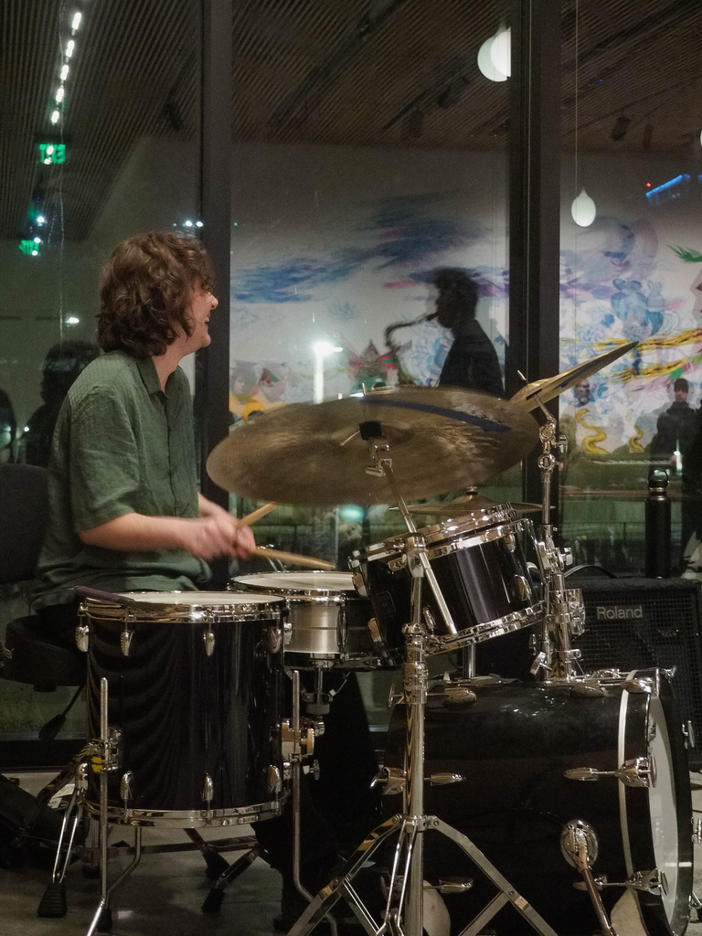 Drummer from both trio groups, smiling while performing and watch the act in front of him.