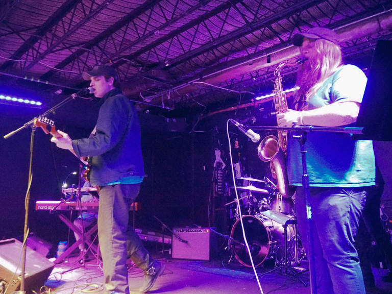 Purple lighting, worms eye view of Dan English on stage singing and playing the guitar. Accompanied by band member playing saxophone.