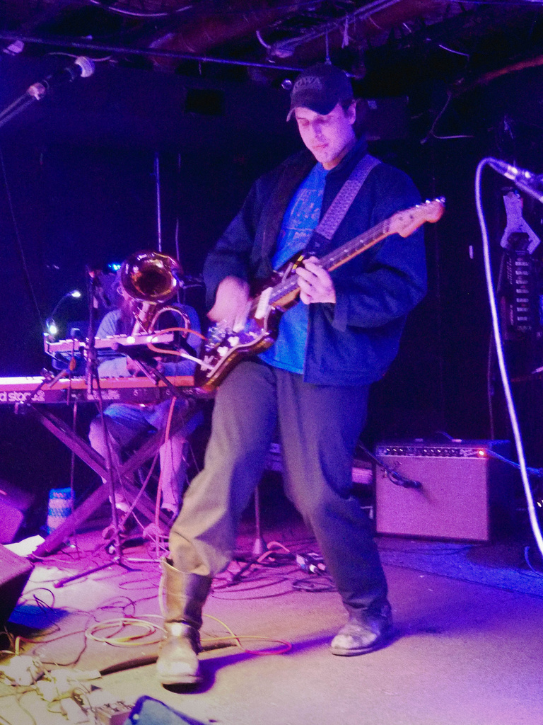 Dan English on stage, vertical shot in blue and purple lighting. Leaning back and playing guitar. He is wearing boots, pants, shirt and jacket with baseball hat.
