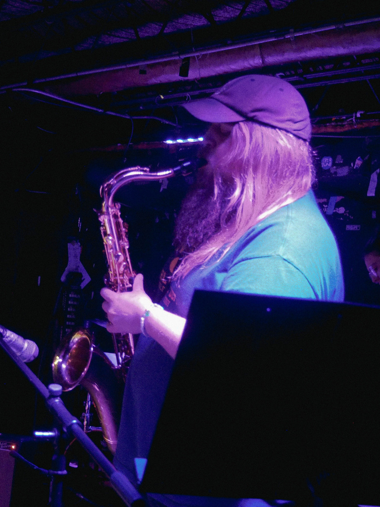 Saxophone player on stage with spotlight. Wearing t-shirt and baseball hat, long-hair and beard.