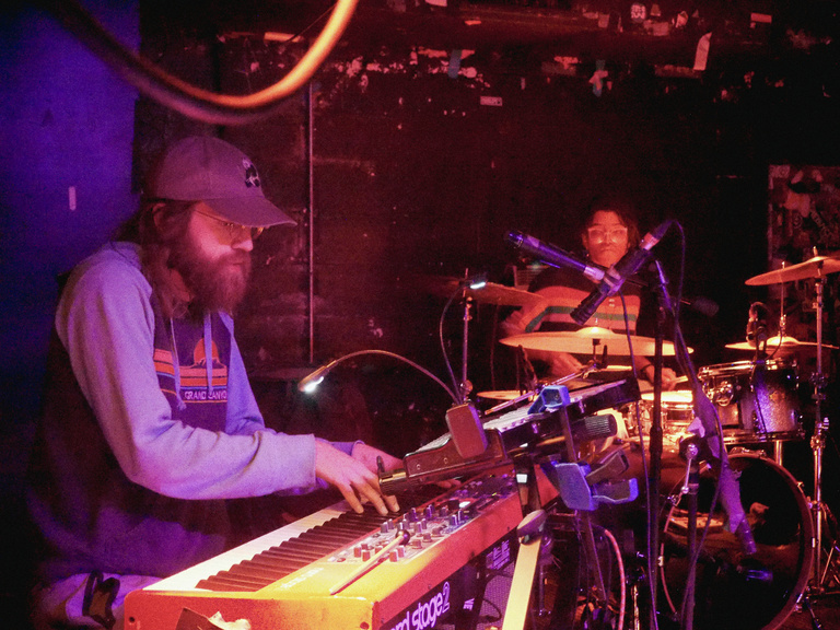 Keys and drummer on stage during a performance. Keys player in foreground, drummer in background. Red lighting and a full stage. 