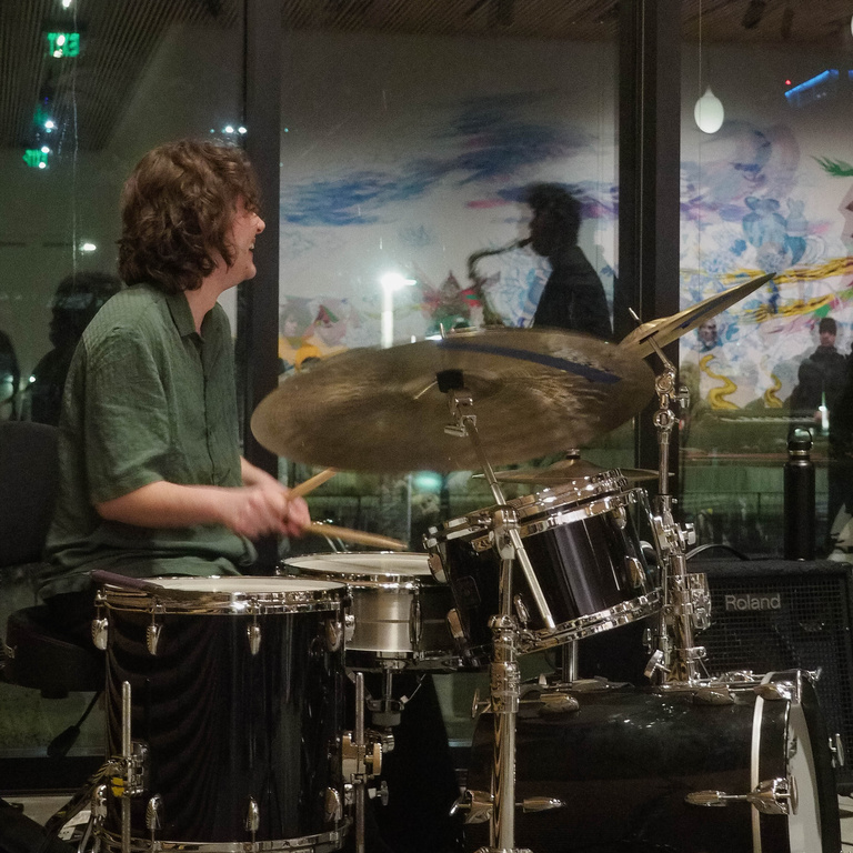 Drummer from both trio groups, smiling while performing and watch the act in front of him.